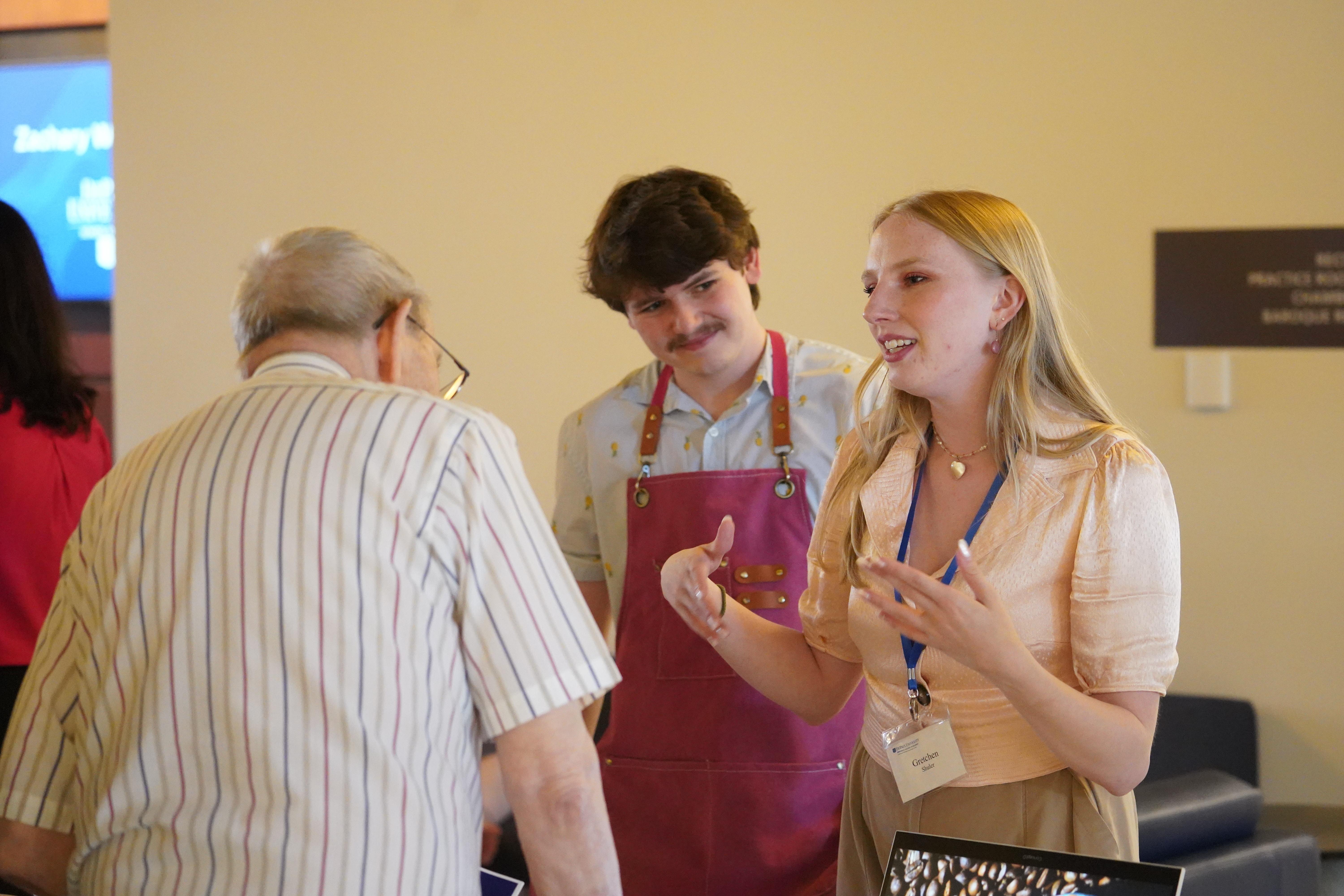 A young woman in the midst of an animated discussion with a visitor to her table; a young man wearing a pink apron stands behind her, nodding approvingly. 